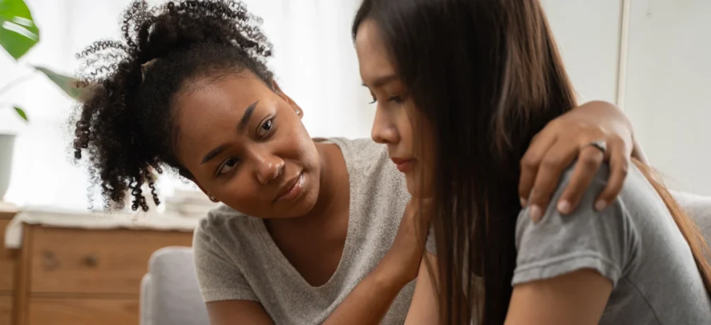 A woman offers comfort to another woman by placing a hand on her shoulder.